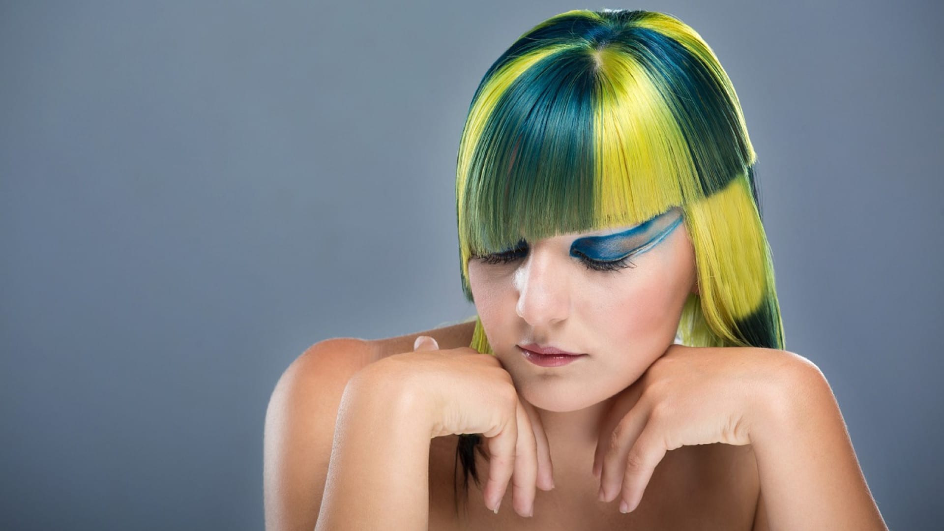 Une personne avec une coiffure colorée avec des nuances de vert et de jaune, grâce à la coloration végétale et un fard à paupières bleu vif, regarde vers le bas avec une expression contemplative.