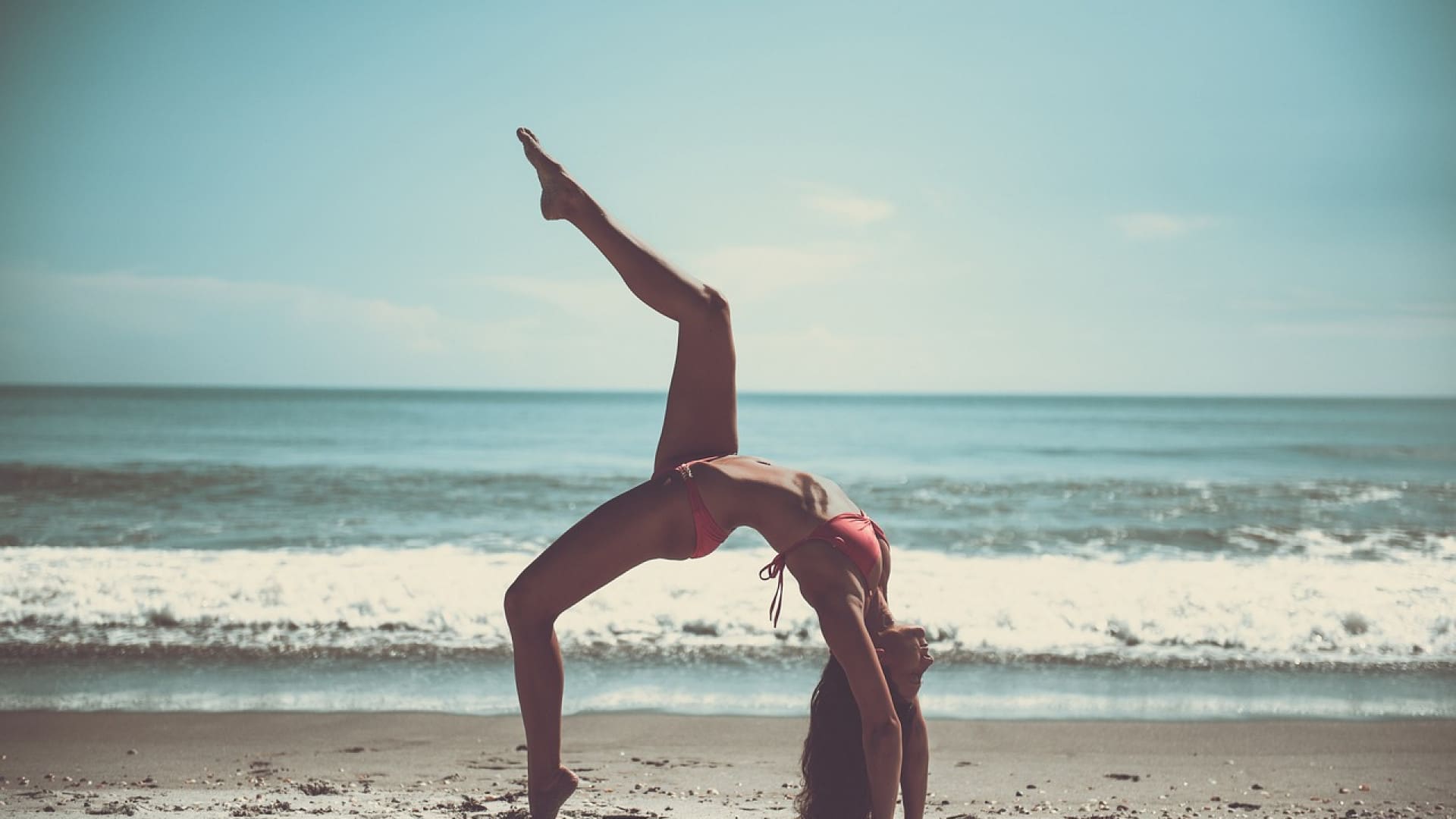 Une personne pratiquant un yoga en faisant un pont sur une plage avec l'océan en arrière-plan.