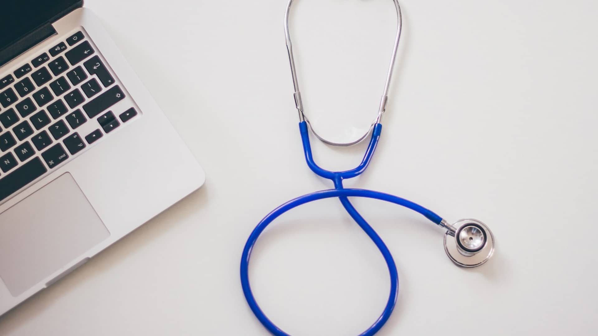 Un ordinateur portable et un stéthoscope sur une surface blanche, symbolisant la santé et la technologie.