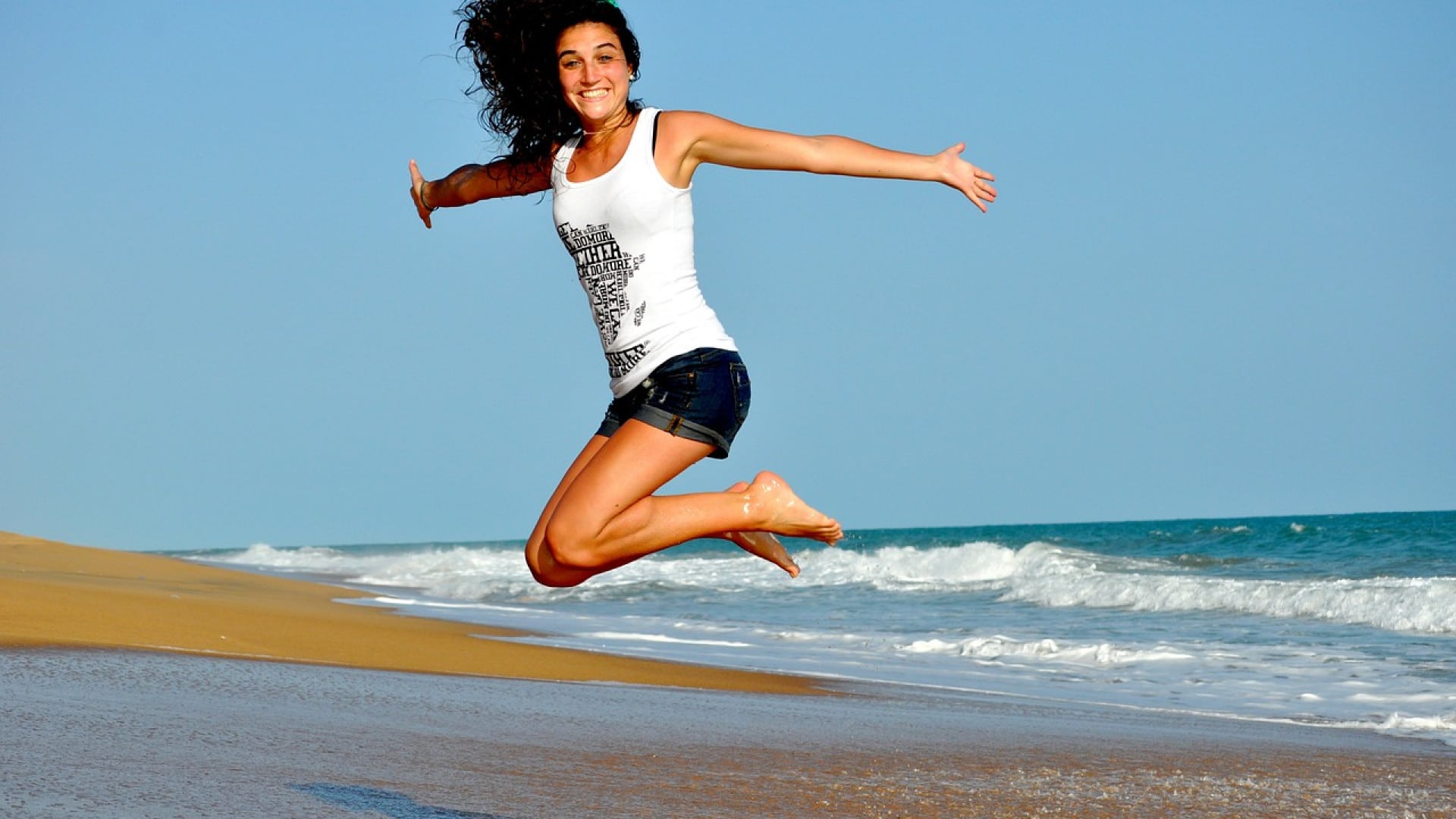 Une femme sautant joyeusement sur une plage de sable fin, prenant soin d’adopter la nouvelle façon de santé avec l’océan en arrière-plan.