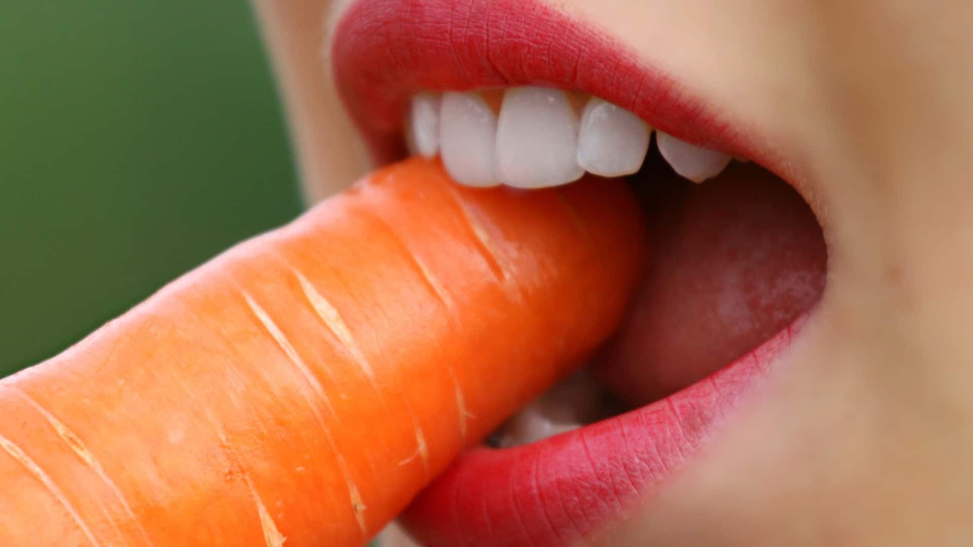Gros plan d'une personne mordant dans une carotte, affichant un sourire radieux.