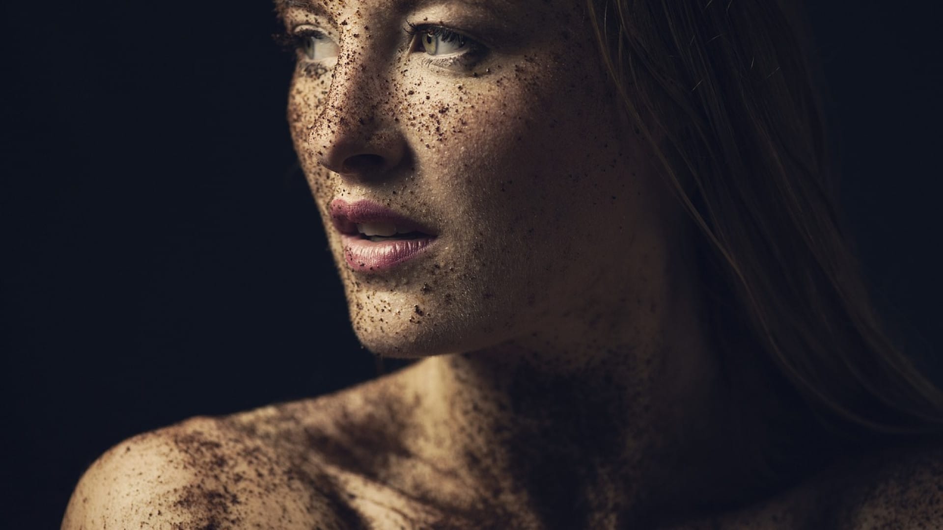 Profil d'une femme avec des taches de rousseur regardant au loin sur un fond sombre, sa peau douce s'éclairant doucement en contraste.
