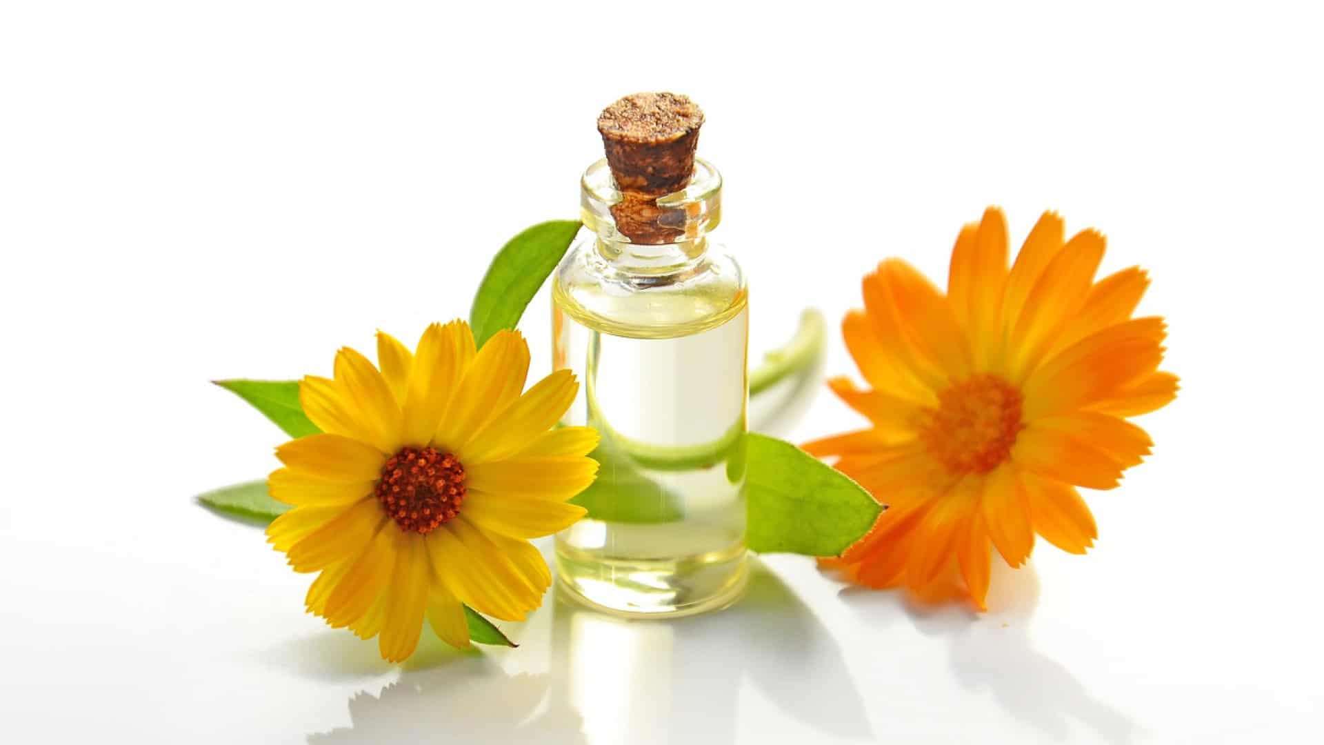 Une petite bouteille en verre contenant de l'huile essentielle pour les soins de la peau, flanquée de deux fleurs jaunes et orange vifs sur fond blanc.