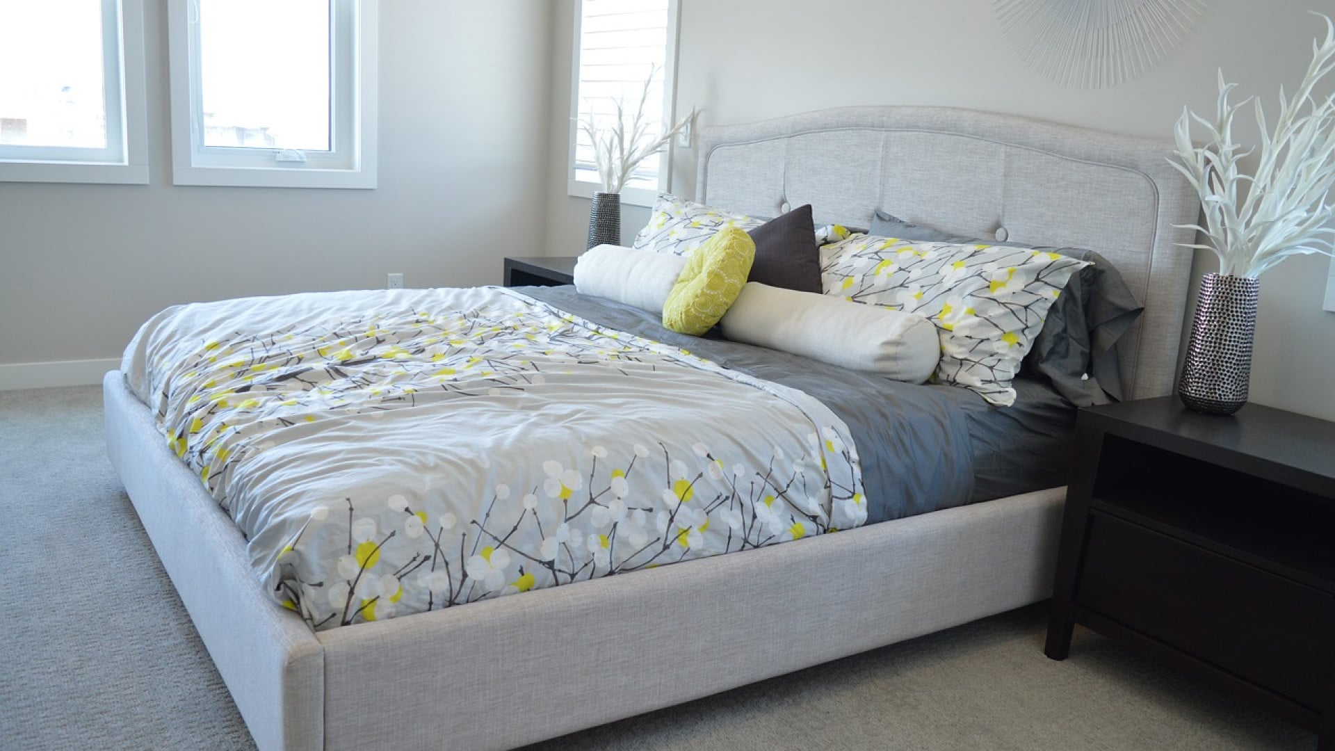 Chambre moderne avec un lit soigneusement fait comprenant un ensemble de literie gris et jaune et des oreillers décoratifs, flanqué d'une table de nuit et d'un vase décoratif.
