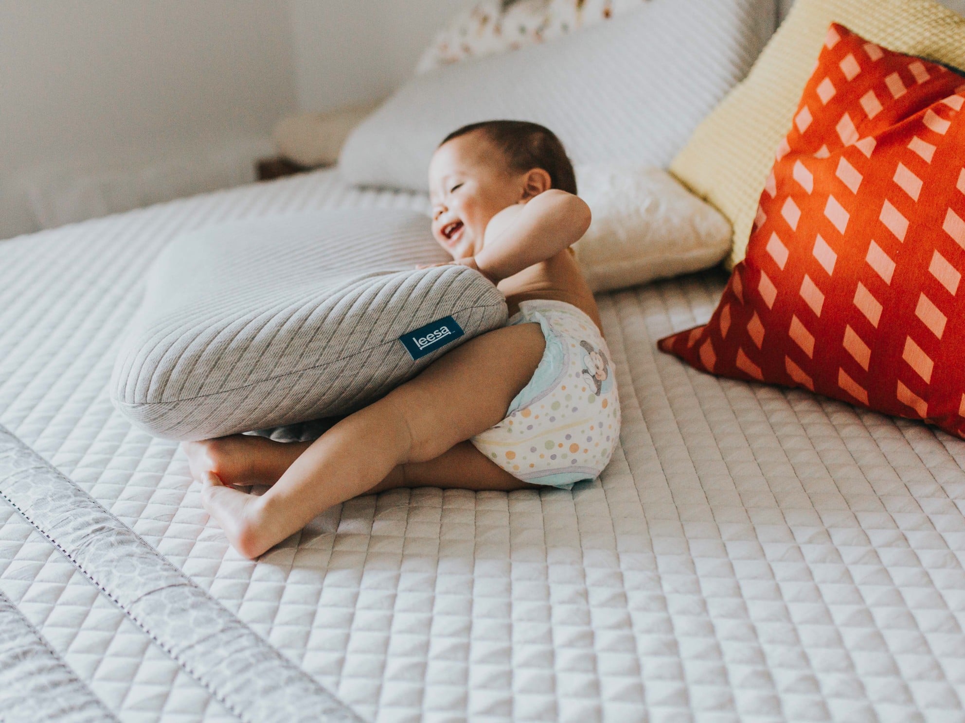 Un bébé en couche est allongé sur un lit, tenant de manière ludique un article de literie avec une expression ravie.