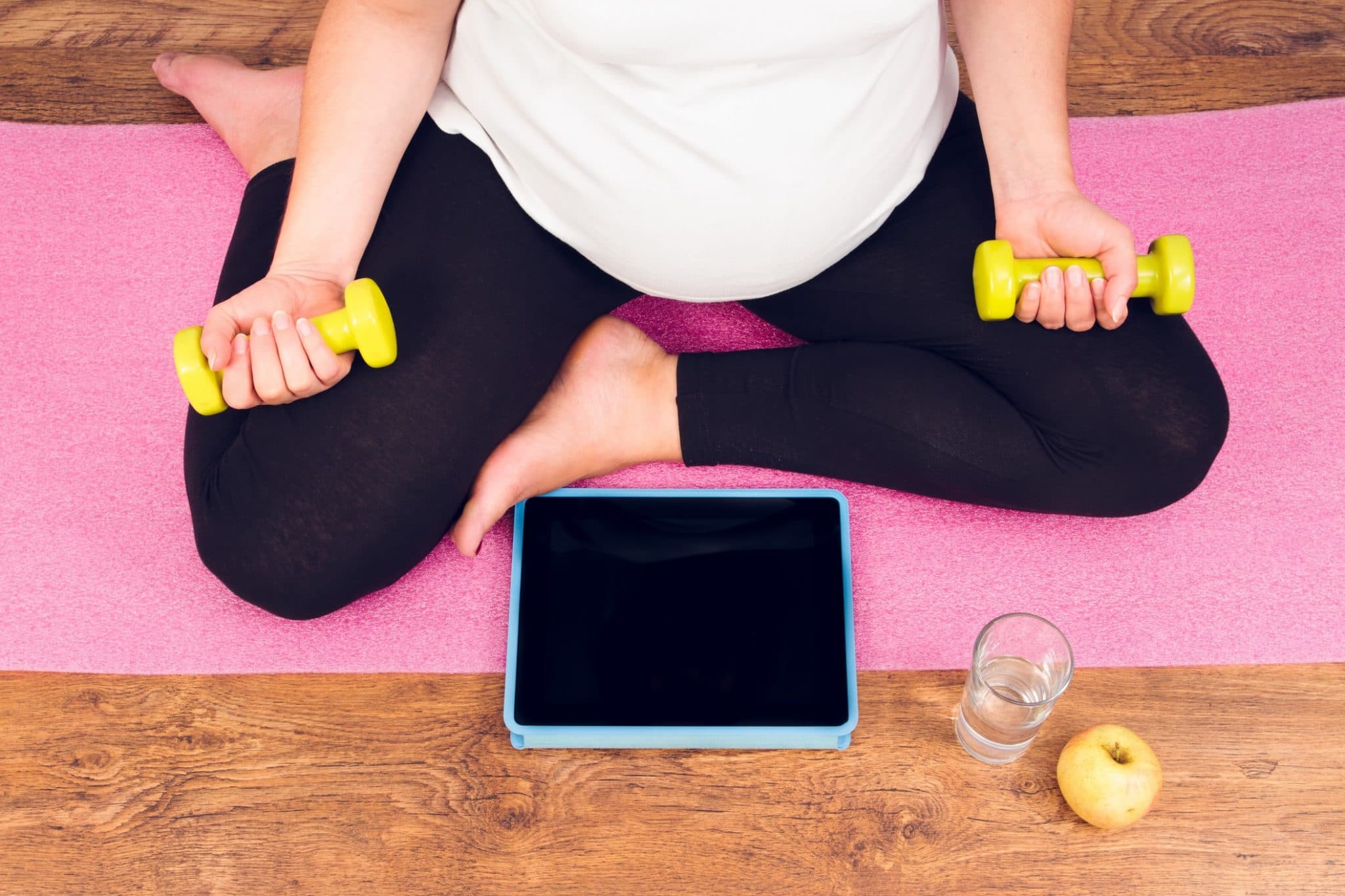 Femme enceinte assise sur un tapis de yoga avec des haltères, une tablette, un verre d'eau et une pomme.