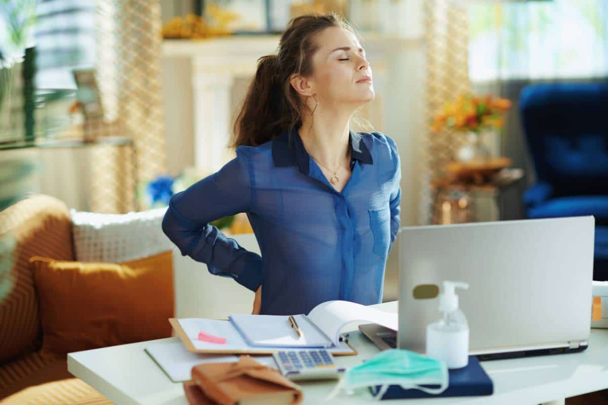 Femme en chemise bleue s'étirant le dos pour mettre fin aux Douleurs aux lombaires sur un poste de travail à domicile avec un ordinateur portable, un ordinateur portable et des articles de sécurité sanitaire.