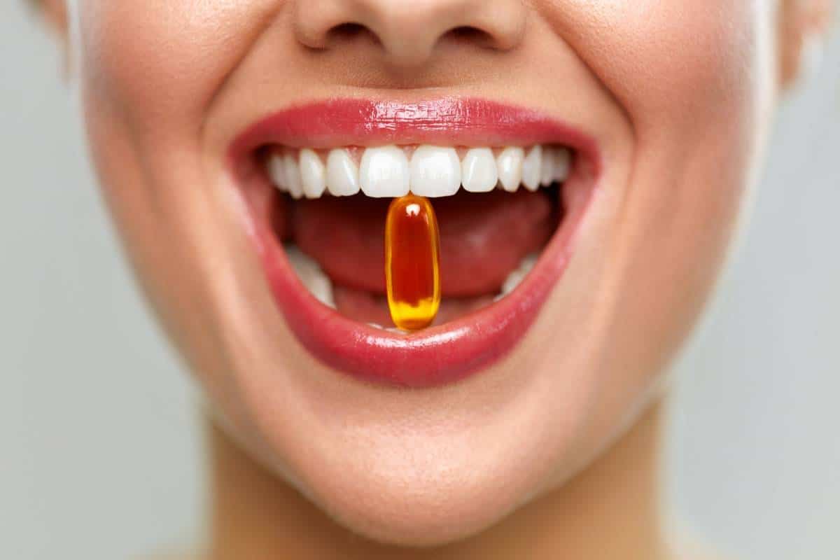 Gros plan d'une personne souriante avec une seule capsule d'oméga-3 équilibrée entre ses dents, tenant un complément alimentaire contre UV.