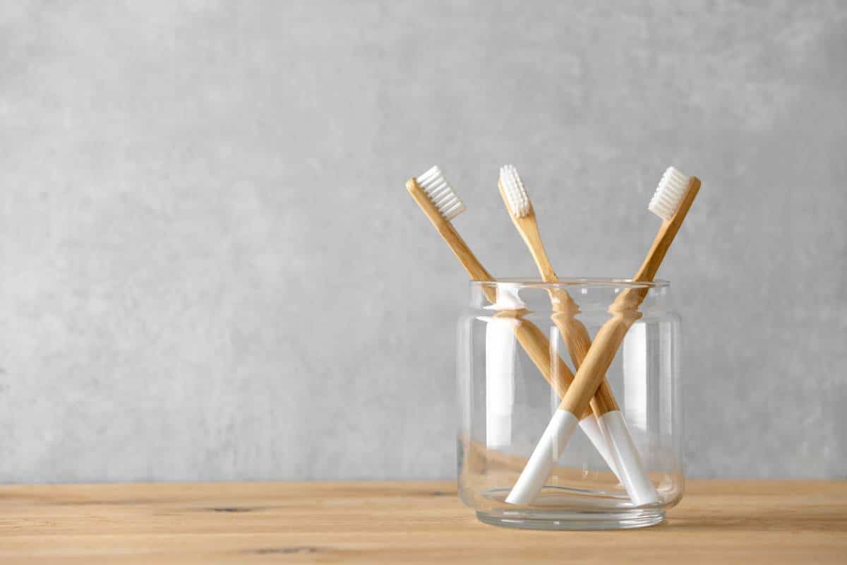 Quatre brosses à dents en bambou dans un bocal en verre sur une surface en bois sur fond gris.