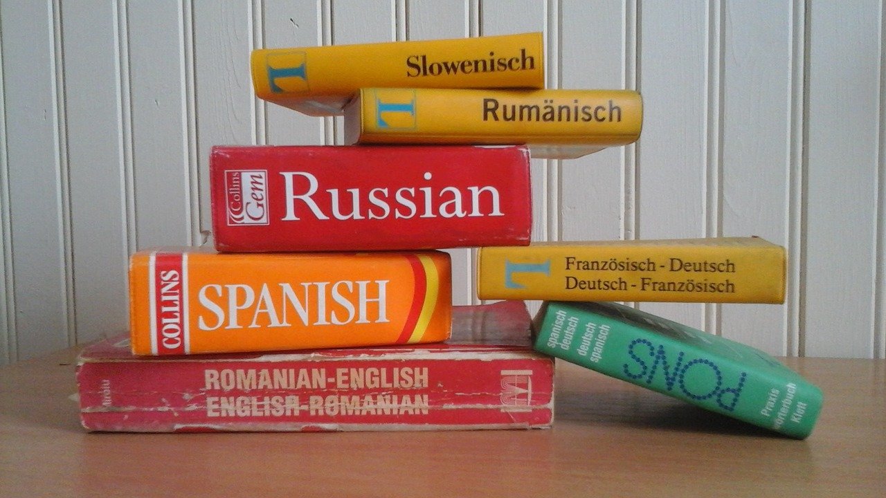 Une pile de dictionnaires de langue pour le slovène, le roumain, le russe, l'espagnol et le français sur une surface en bois contre un mur rayé.