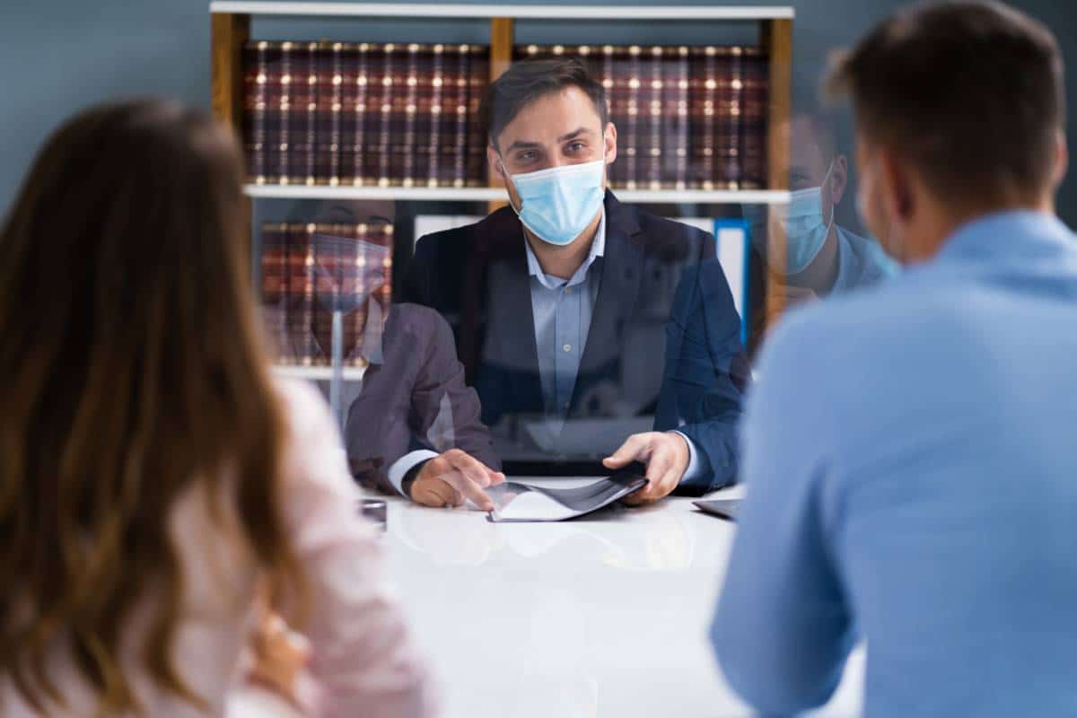 Un professionnel portant un masque facial est assis derrière une barrière de protection transparente lors d'une réunion avec deux clients.