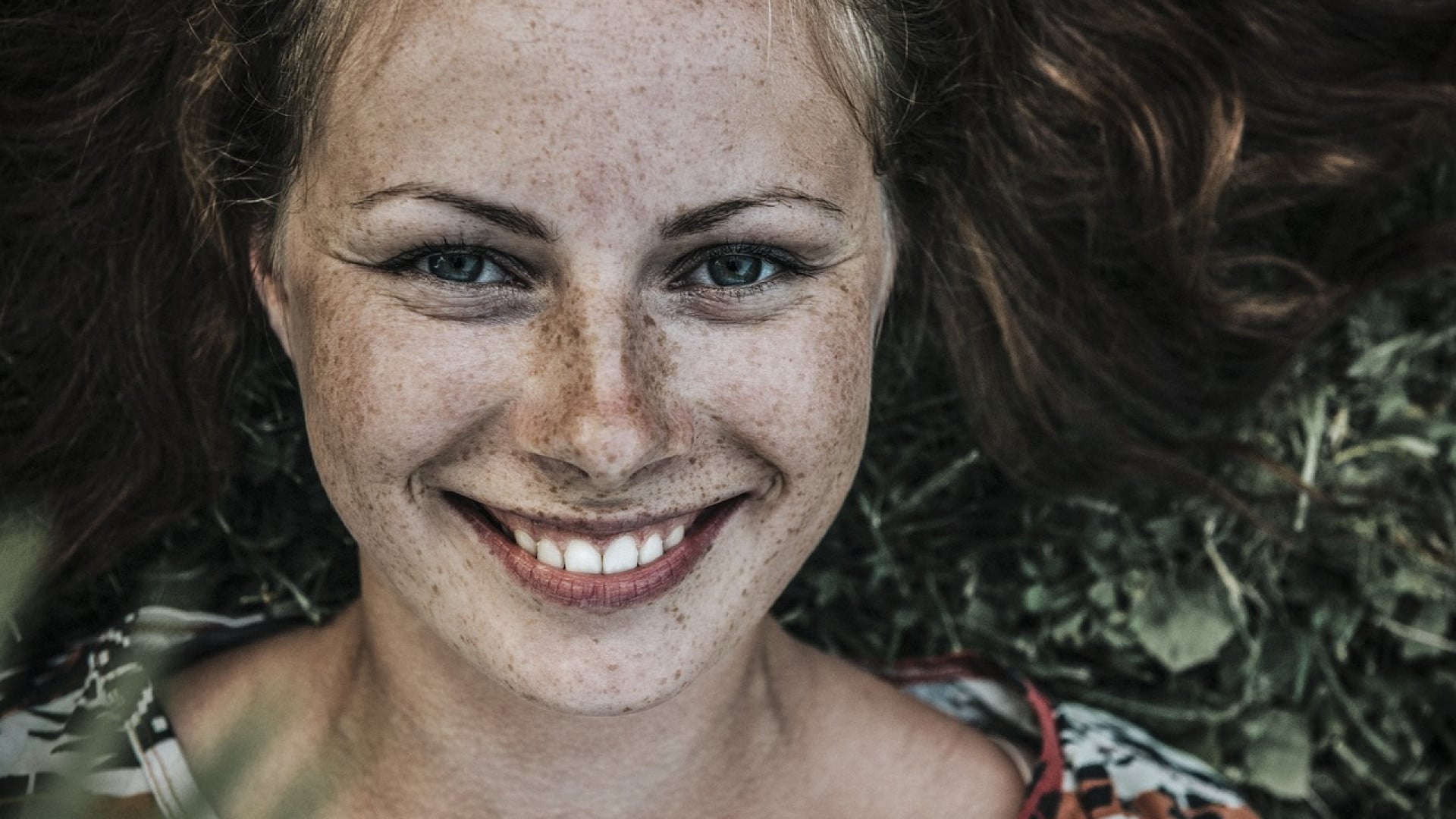 Gros plan d'une femme souriante avec des taches de rousseur allongée sur l'herbe.
