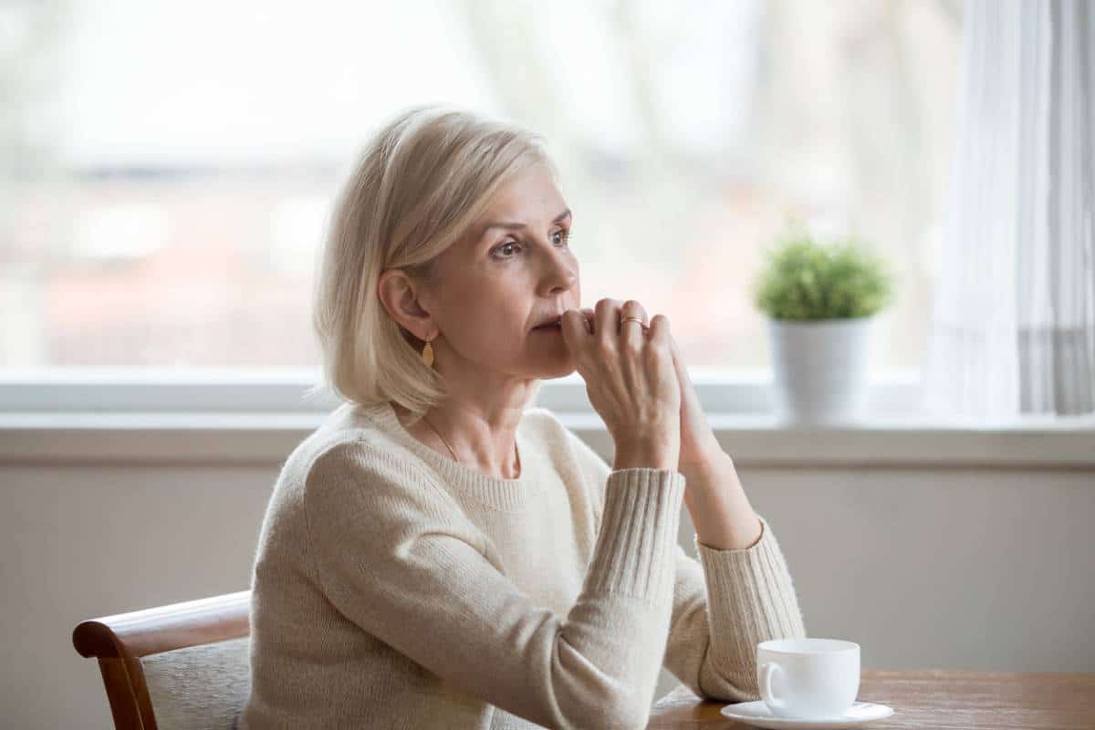 Une femme pensive assise à une table avec une tasse de café, regardant par la fenêtre, prête à poser des questions.