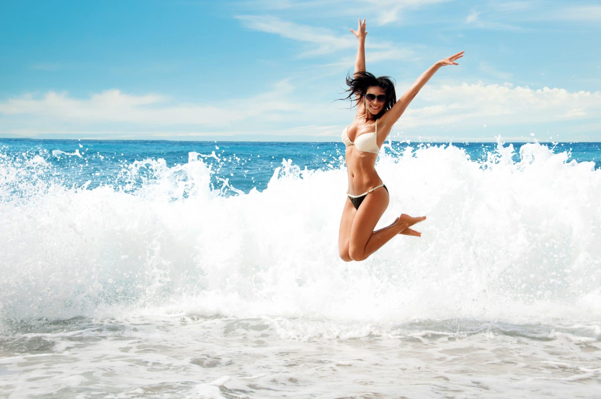 Femme sautant joyeusement sur la plage avec des vagues se brisant en arrière-plan, incarnant la liberté de se baigner avec ses règles.