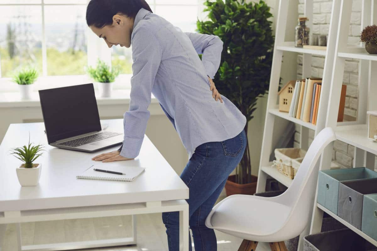 Femme souffrant de maux de dos alors qu'elle se tient debout à un poste de travail avec un ordinateur portable.