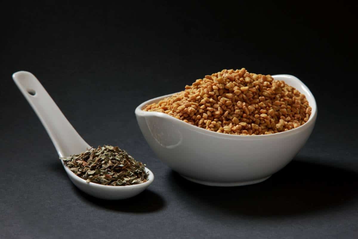 Un bol de graines de sésame à côté d'une cuillerée d'herbes mélangées et d'un petit flacon d'huile essentielle sur fond sombre.