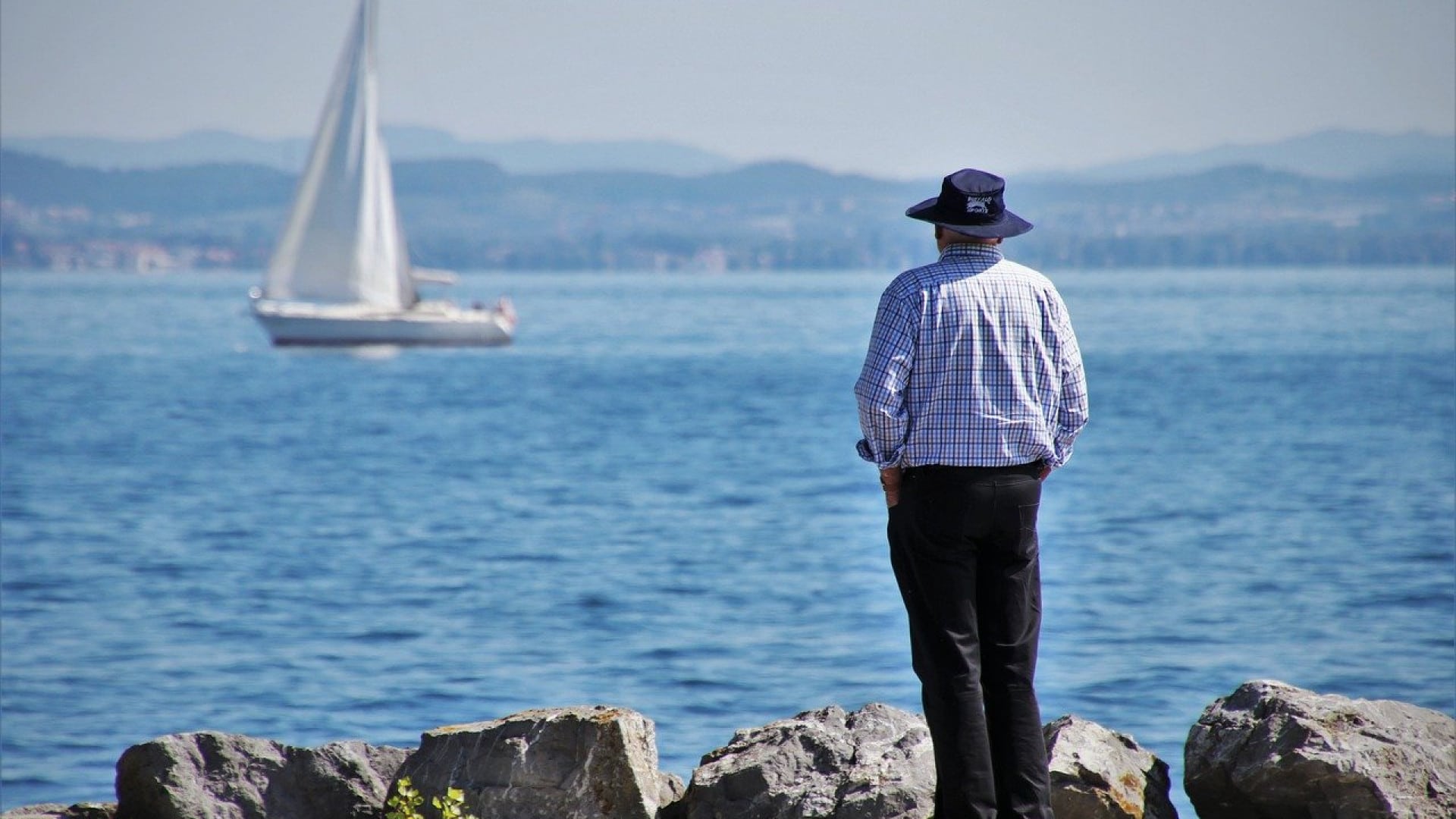 Homme au chapeau debout sur des rochers et observant un voilier sur l'eau.