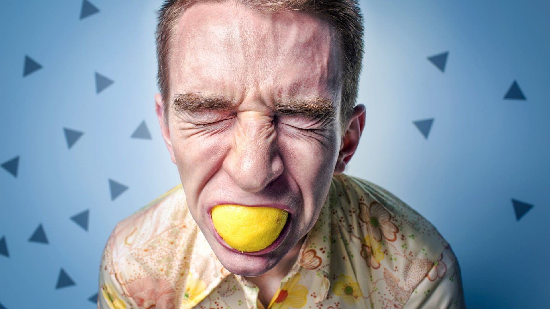 Homme grimaçant en mordant un citron sur un fond bleu avec des motifs triangulaires.