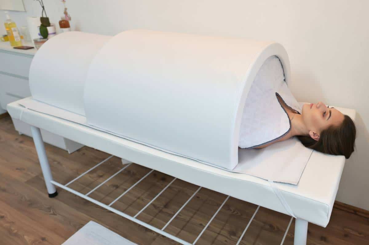 Femme allongée à l'intérieur d'une capsule blanche ouverte pour un soin de relaxation ou de sauna japonais.