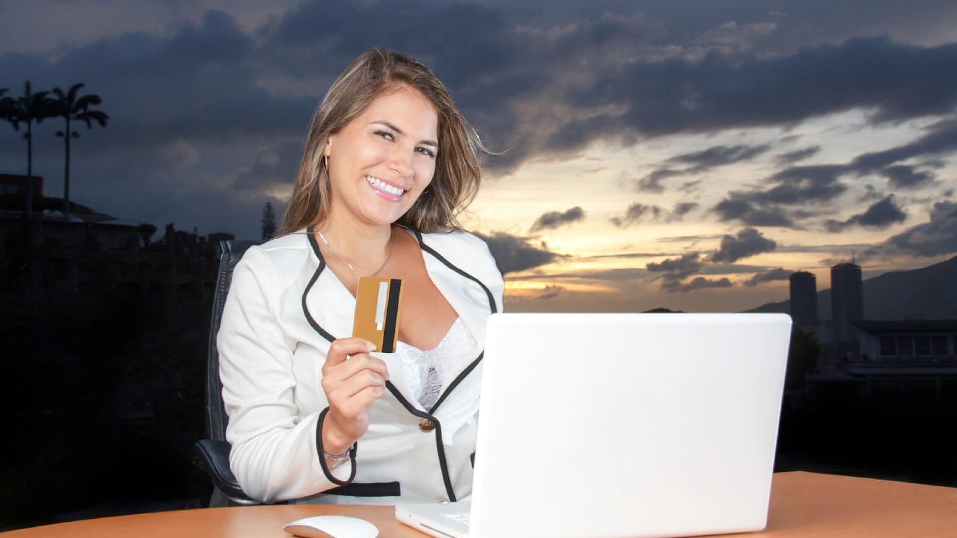 Femme souriante avec une carte de crédit assise devant un ordinateur portable à l'extérieur au crépuscule.