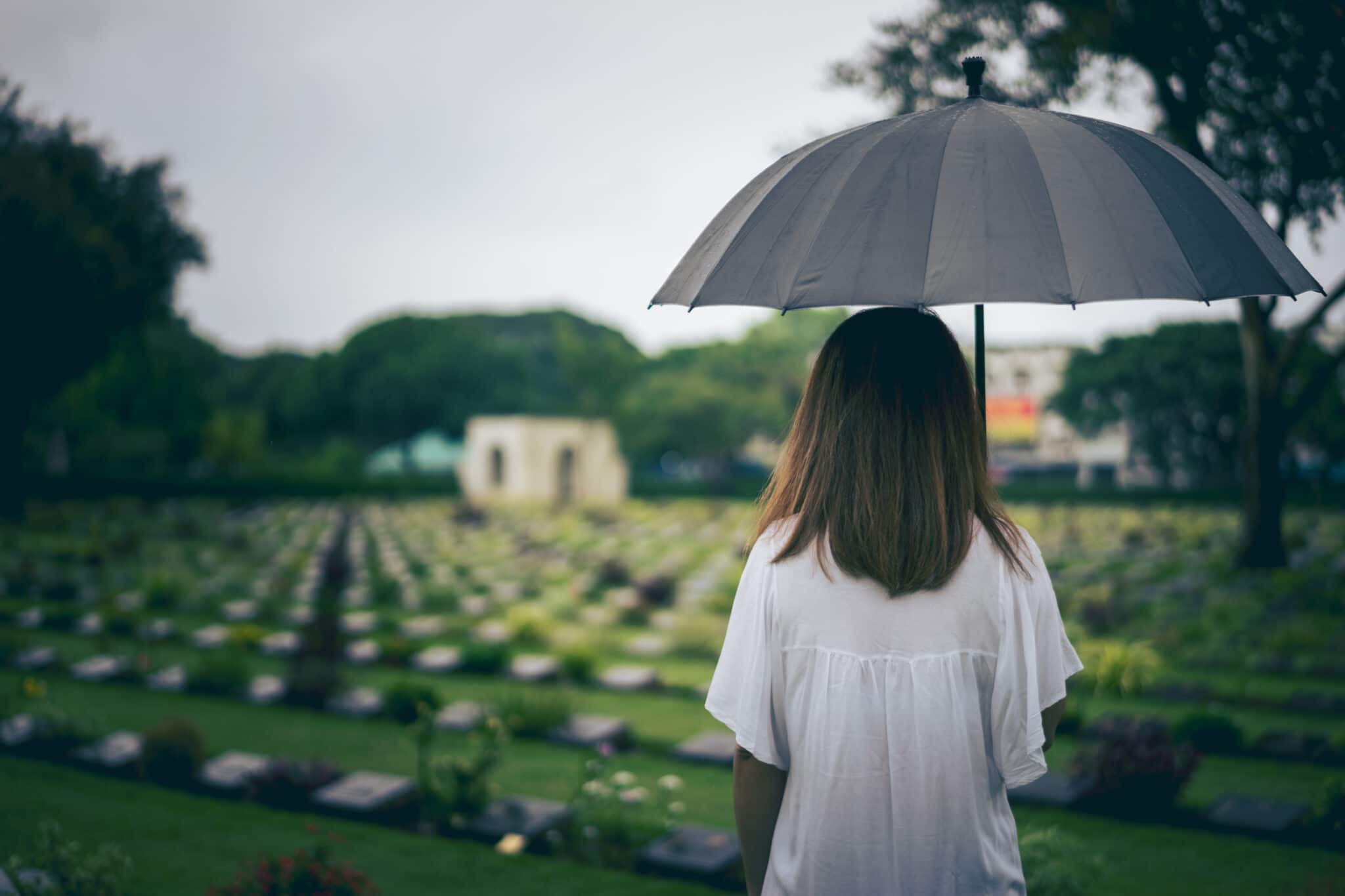 Une personne tenant un parapluie se tient devant un cimetière par temps couvert, incarnant le deuil solennel de la mort d'un proche.