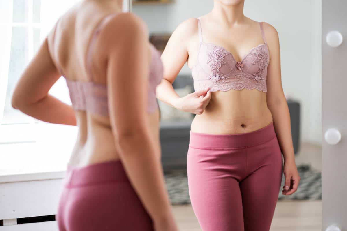 Femme en soutien-gorge et leggings regardant son reflet dans le miroir après une intervention d'augmentation mammaire.