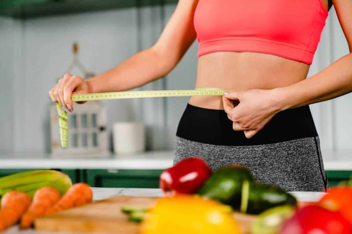 Une personne mesurant sa taille avec un ruban à mesurer dans une cuisine avec des légumes frais au premier plan.