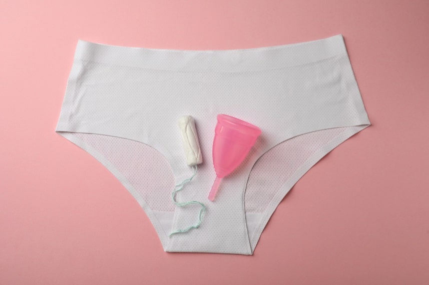 Une paire de sous-vêtements blancs avec un tampon, une coupe menstruelle et une serviette hygiénique placée dessus sur fond rose.