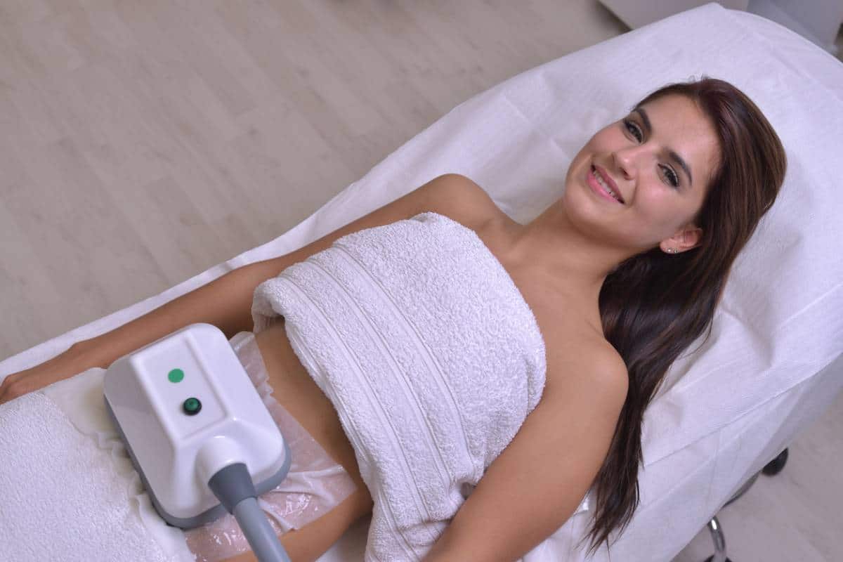 Une femme souriante allongée sur un lit de spa avec un appareil de modelage du corps placé sur son abdomen.