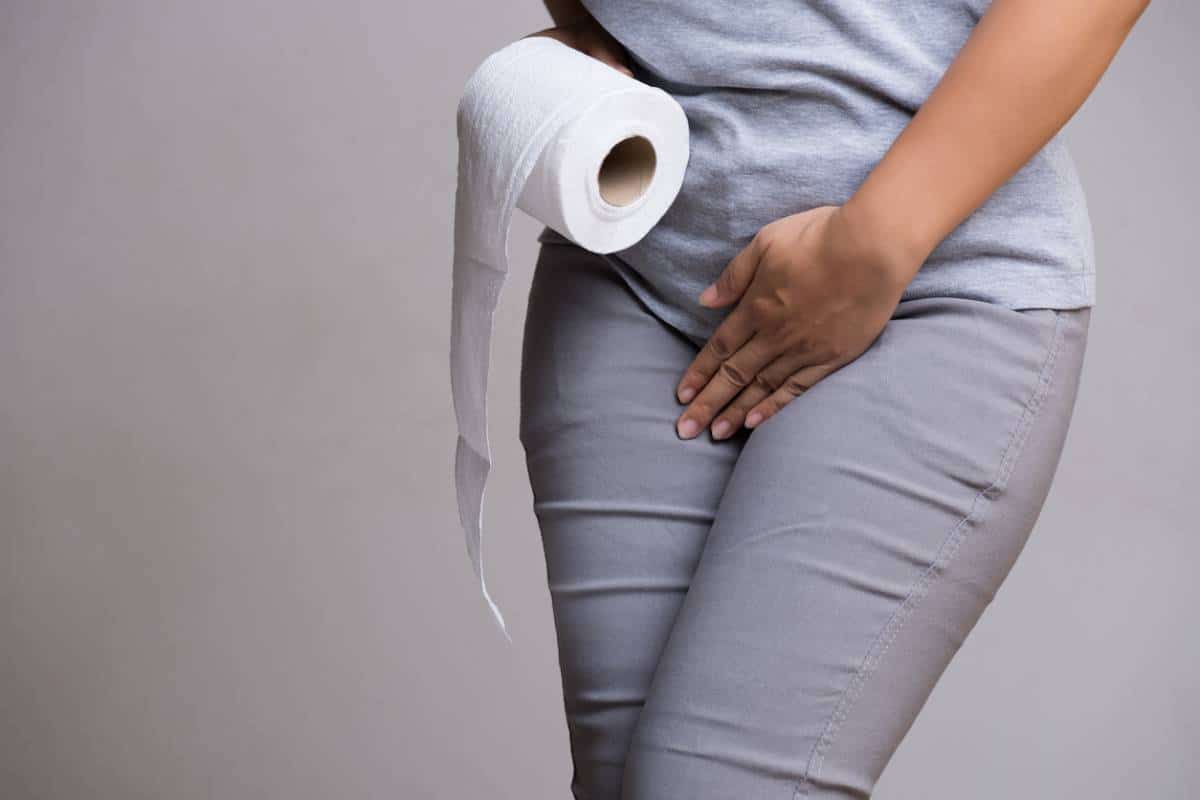 Une personne tenant un rouleau de papier toilette tout en appuyant son autre main contre le bas de son dos, suggérant un inconfort possiblement lié à des fuites urinaires.