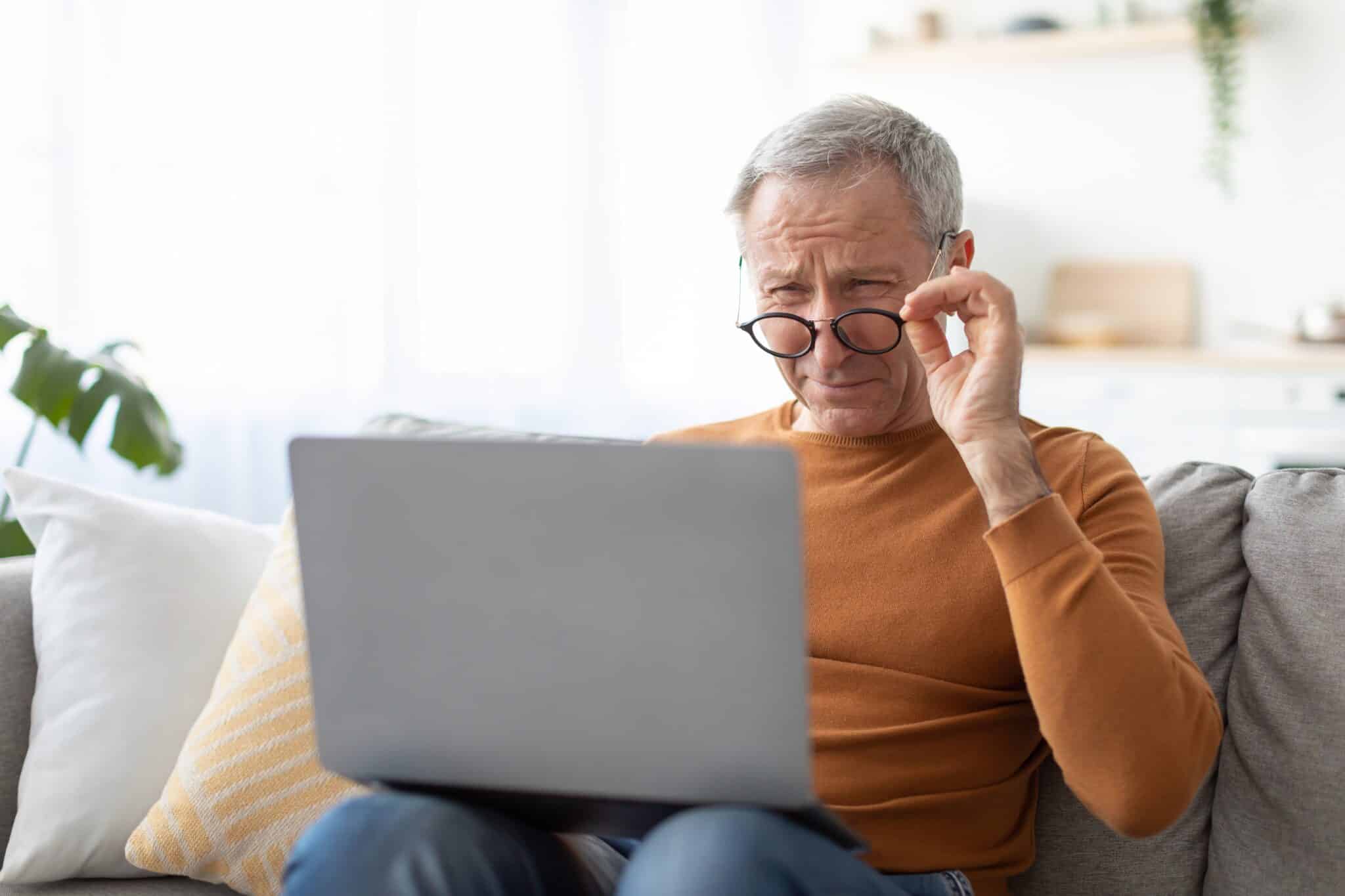 Homme âgé ajustant ses lunettes tout en regardant l'écran d'un ordinateur portable avec une expression inquiète en raison de la dégénérescence maculaire liée à l'âge (DMLA).