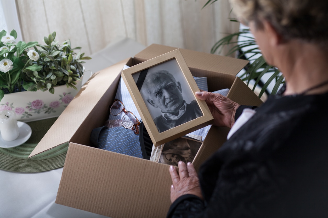 Une personne âgée tenant une photo encadrée de son conjoint tout en regardant dans une boîte d'objets personnels, alors qu'elle traverse le processus de deuil après le décès de son partenaire.