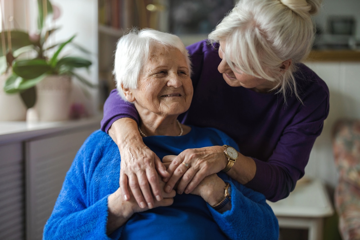 Une femme plus jeune, fournissant des services d'aide à domicile, embrasse par derrière une femme âgée, toutes deux souriant chaleureusement.