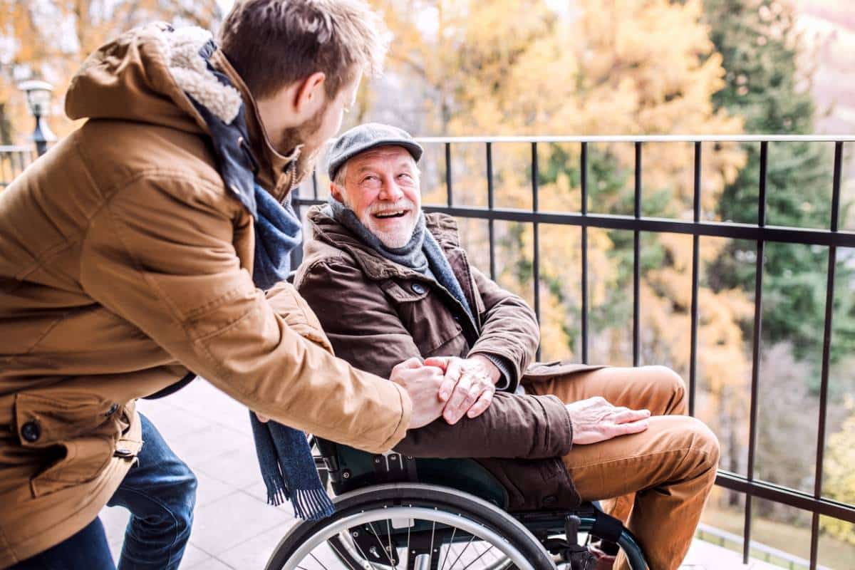 Une jeune personne interactive avec une personne handicapée plus âgée en fauteuil roulant sur un balcon, tous deux souriants.