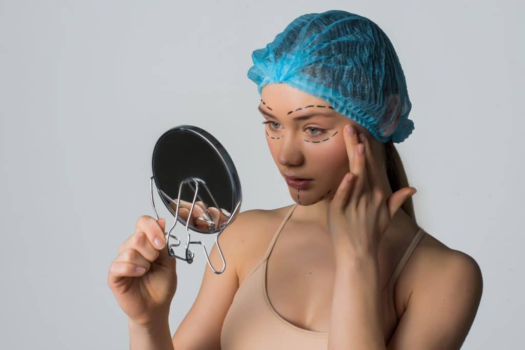 Une femme portant un bonnet chirurgical se regarde dans un miroir avant ses interventions chirurgicales esthétiques.