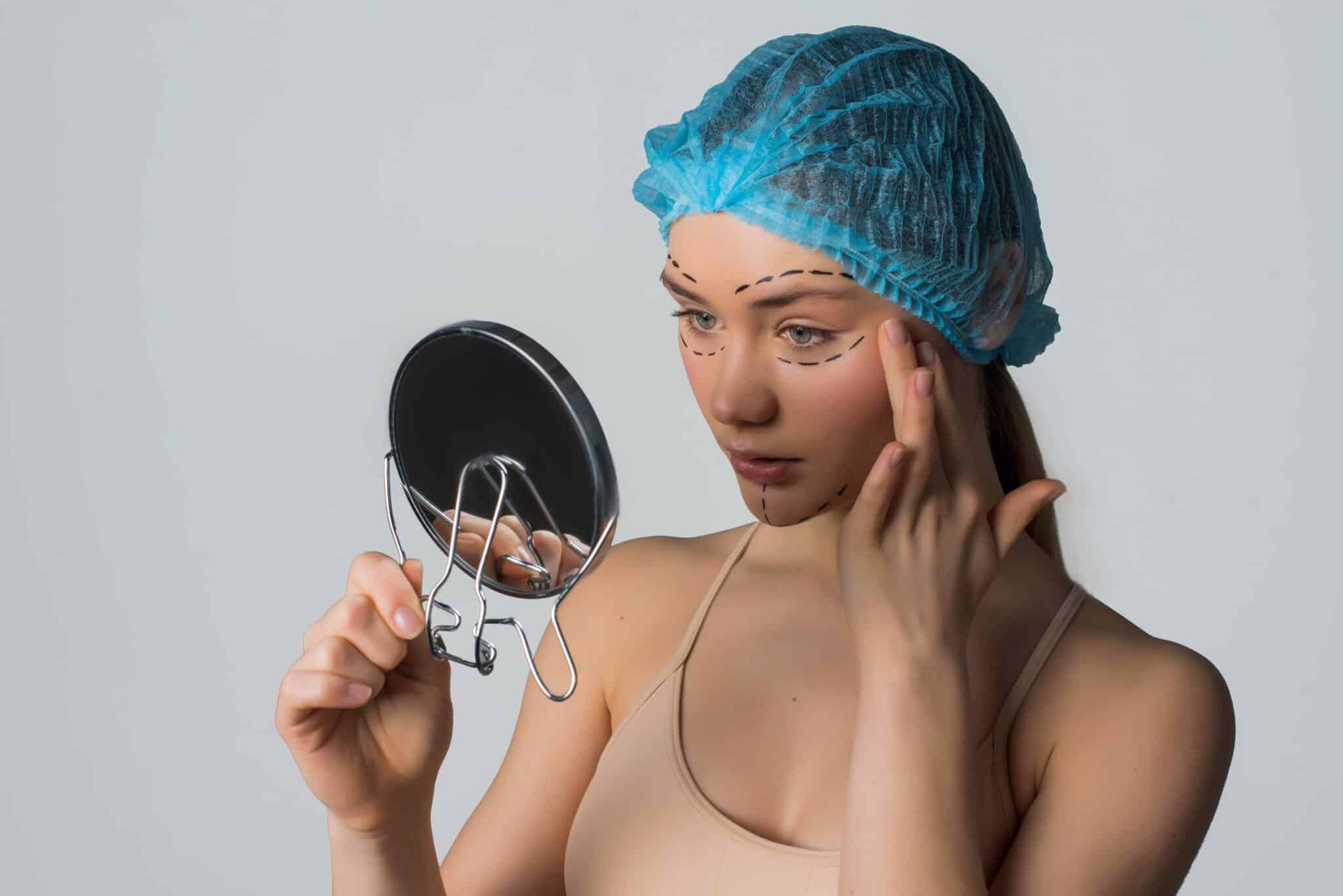 Une femme portant un bonnet chirurgical se regarde dans un miroir avant ses interventions chirurgicales esthétiques.